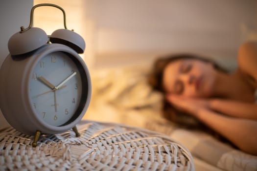 A large alarm clock stands on the cabinet next to the sleeping young woman. A girl under a soft blanket.