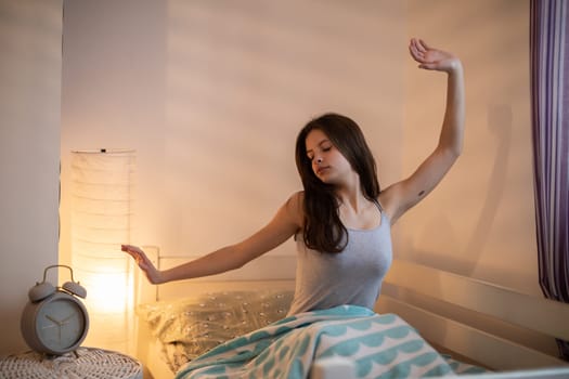The girl drags herself on the bed in the bedroom. A large alarm clock stands on the bedside table.