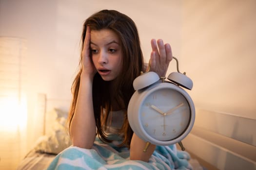 A tired girl holds a bright alarm clock in her hand. The light of a lamp on the wall.