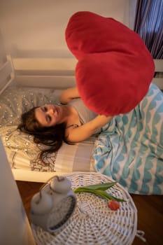 A teenage girl holds a red heart-shaped pillow above her while lying on her bed. The young girl dreams of her first true love.