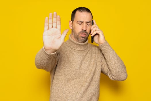 Bearded hispanic man in a turtleneck asking not to be disturbed while talking on his smartphone, he is very pissed off the interruption has taken him out of his mind. Isolated on yellow background