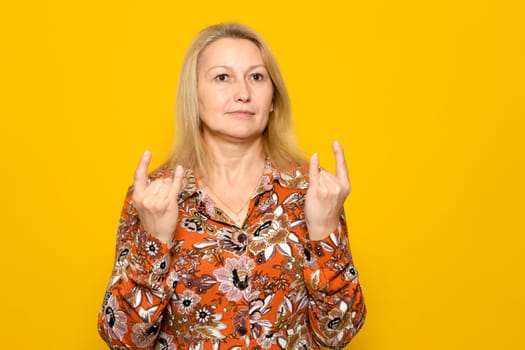 Caucasian blonde woman in a patterned dress making the rock gesture with both hands, isolated on yellow studio background. Concept of rebellion.