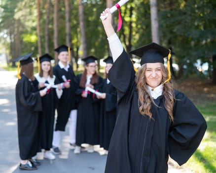 Group of happy students in graduation gowns outdoors. A young girl with a diploma in her hands in the foreground