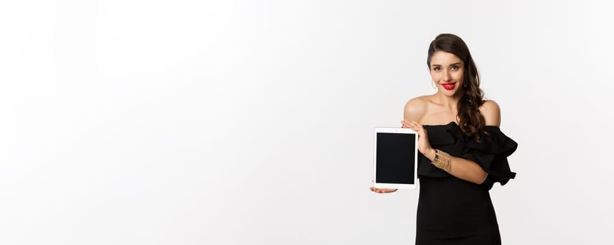 Online shopping concept. Tempted pretty woman in black dress showing digital tablet screen, standing over white background. Copy space