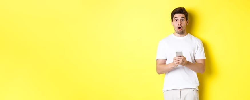 Man looking surprised, using smartphone, open mouth and saying wow, standing against yellow background. Copy space