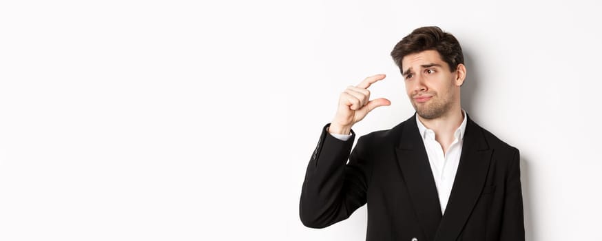 Close-up of handsome businessman in trendy suit, showing something small with disappointment, standing against white background.