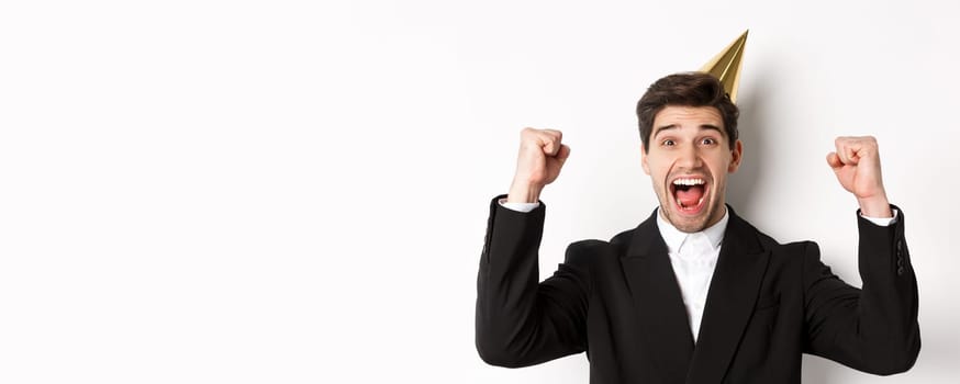 Close-up of happy good-looking man, wearing party hat and suit, raising hands up and rejoicing, celebrating new year, standing against white background.