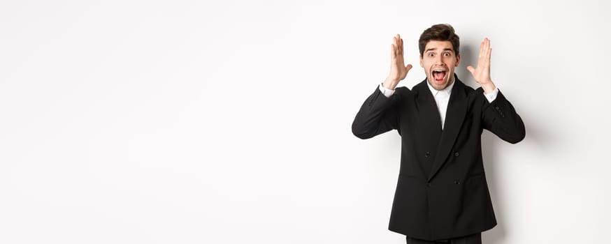 Frustrated and concerned man in black suit, screaming in panic and looking at something shocking, standing over white background.