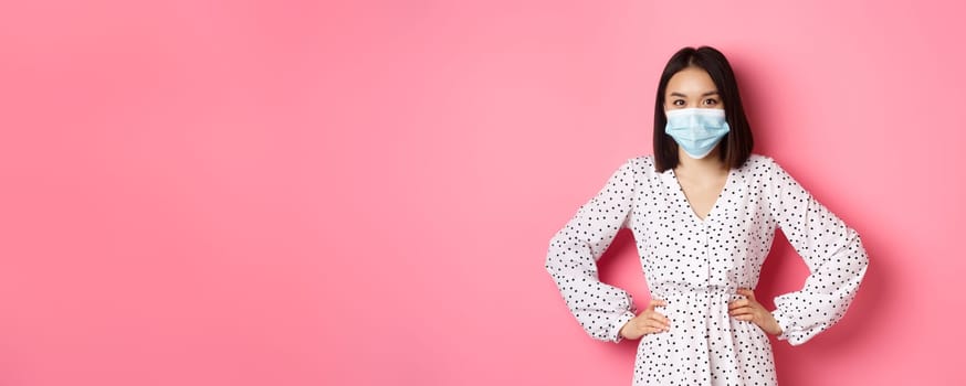 Covid-19, quarantine and lifestyle concept. Cute asian woman wearing medical mask and dress, protect herself from coronavirus, standing over pink background.