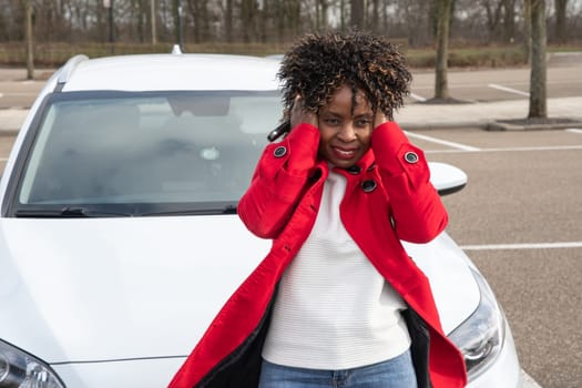 frustrated african american woman holding her hair with her hands due to strong wind standing in front of her white car, High quality photo
