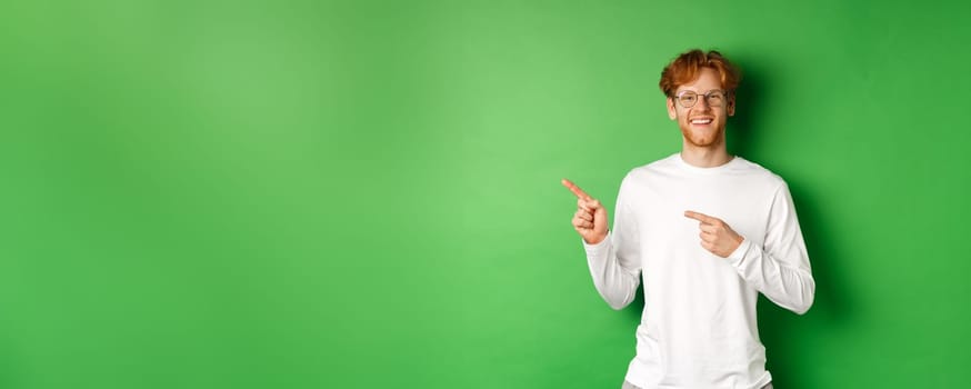 Handsome young man with red hair and beard smiling, pointing fingers left at logo, showing advertisement, standing over green background.