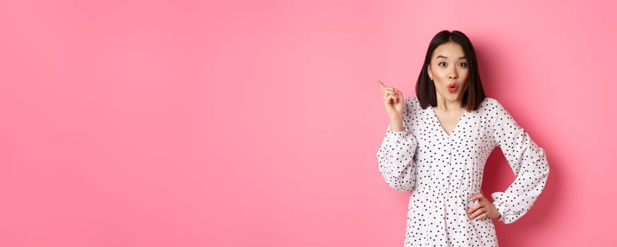 Excited asian female model showing promo offer, pointing at upper left corner and staring at camera amazed, wearing trendy spring dress, standing over pink background.