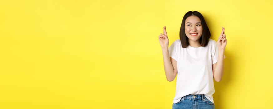 Hopeful asian girl smiling optimistic, feeling lucky, cross fingers and making wish, standing over yellow background.