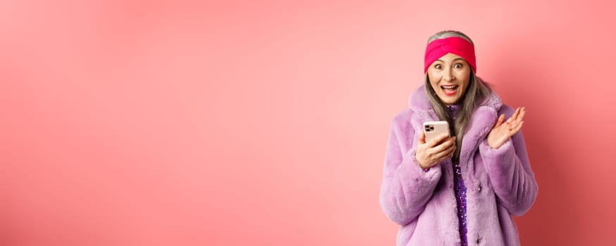 Online shopping and fashion concept. Asian mature woman looking excited as checking out internet promo offer on smartphone, looking happy at camera, pink background.