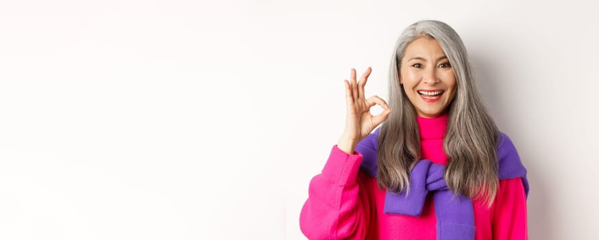 Close up of beautiful asian mature woman showing OK sign and smiling, approve and like product, standing satisfied against white background.