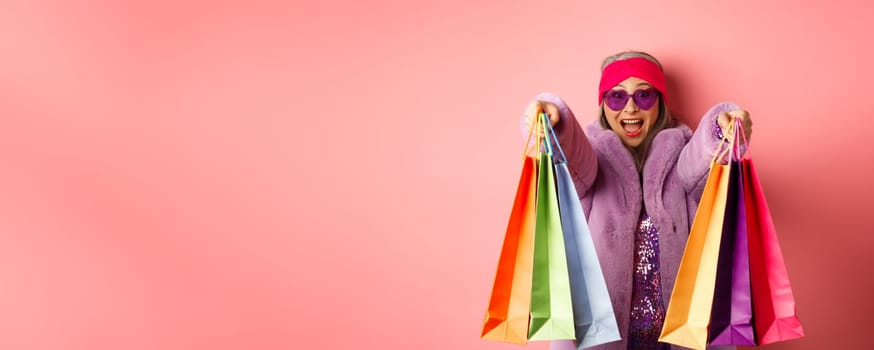 Happy asian adult woman going shopping, extend hands with shop bags and smiling cheerful, standing over pink background.