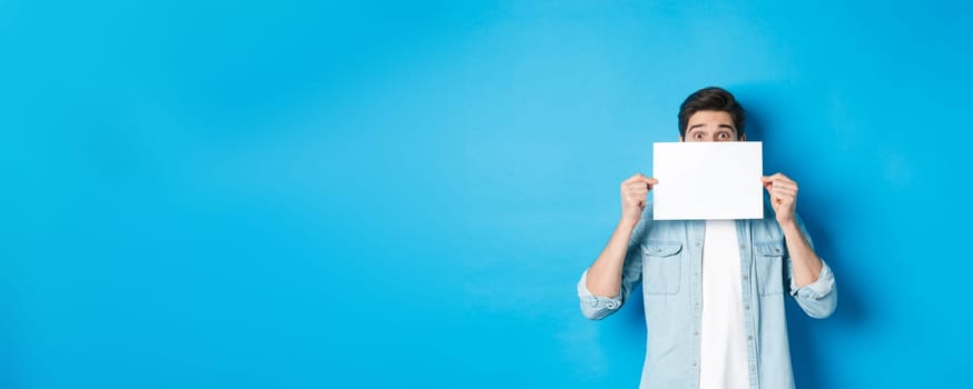 Excited man hiding face behind blank paper, place for your logo, standing over blue background.