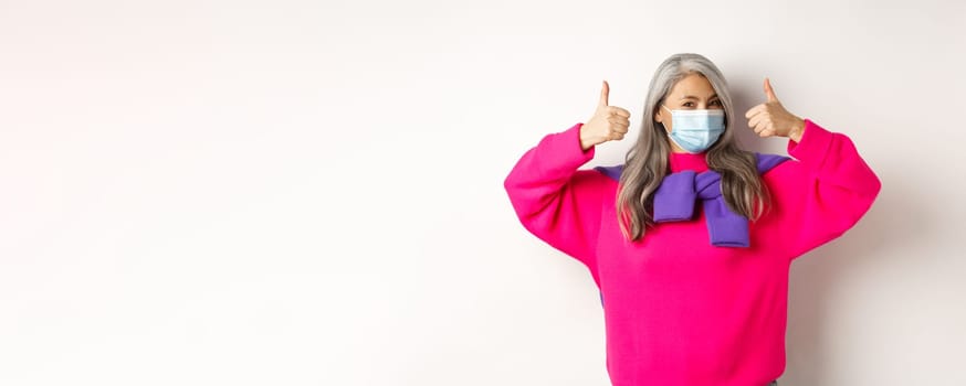 Covid, pandemic and social distancing concept. Cheerful and stylish asian senior woman wearing medical mask and showing thumbs-up, standing over white background.