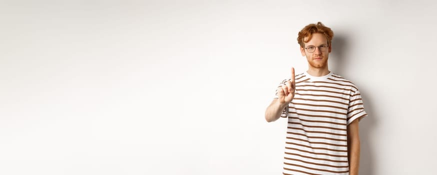 Young man with red hair and glasses shaking finger in prohibition, taboo gesture, disapprove and stop something, standing over white background.