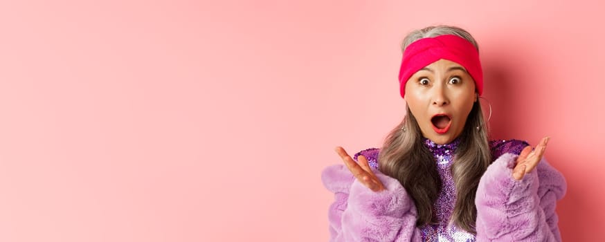 Fashion. Close-up of fashionable asian elderly woman reacting to special offer, scream and gasping surprised at camera, standing over pink background.