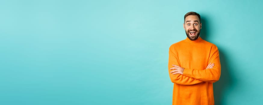 Image of happy and surprised man reacting to news, looking amazed, standing in orange sweater against turquoise background.