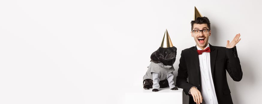Happy young man and cute black dog wearing party cones, celebrating birthday, guy friendly saying hello and waving hand, standing over white background.