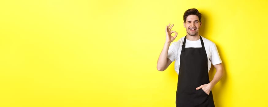 Confident and handsome waiter showing ok sign, wearing black apron and standing against yellow background.