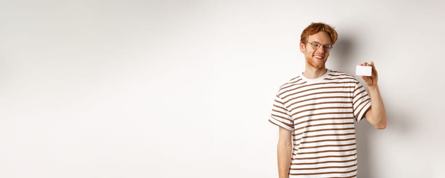 Shopping and finance concept. Cheerful redhead male student in glasses showing plastic credit card and looking satisfied, standing over white background.