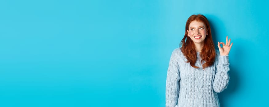 Image of beautiful redhead woman showing okay sign and smiling satisfied, like and agree, standing against blue background.