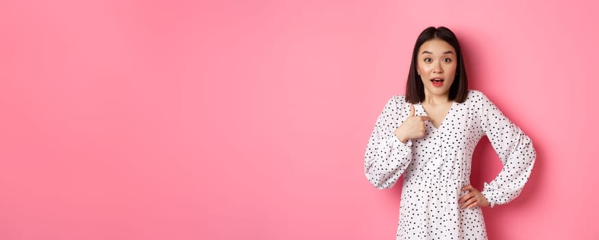 Beautiful brunette asian woman pointing finger at camera, looking surprised as being chosen, standing in dress against pink background.
