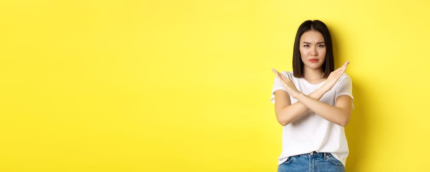 Serious and assertive korean woman showing cross stop gesture, frowning and telling no, prohibit action, disapprove something bad, standing over yellow background.