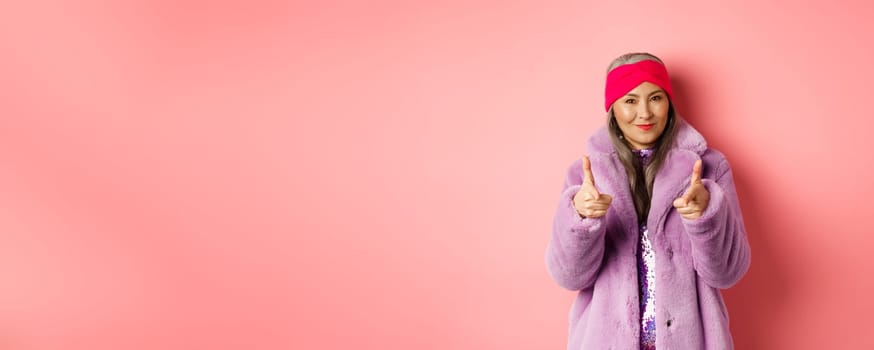 Fashion and shopping concept. Cool asian senior woman in stylish fake fur coat pointing fingers at camera, asking you to check out promo offer, standing over pink background.