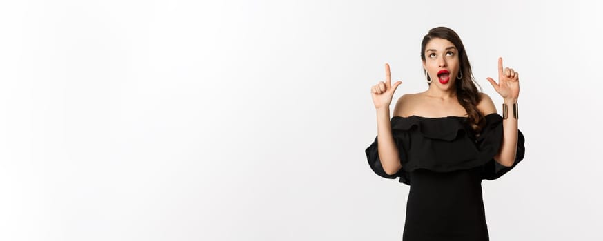 Fashion and beauty. Amused woman in black dress looking and pointing fingers up at promo offer, standing over white background.