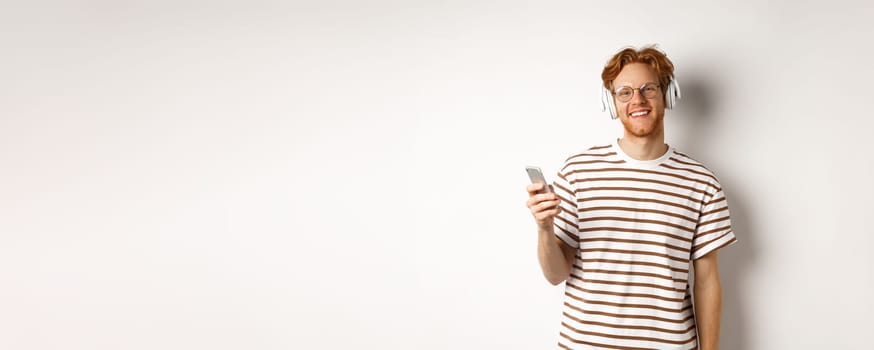 Technology concept. Young man with red hair and beard listening music in headphones and using smartphone, smiling at camera, white background.