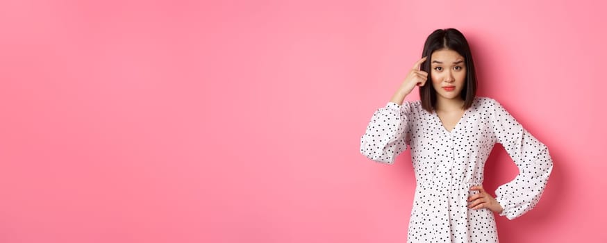 Image of attractive asian lady scolding person, pointing at head and staring at camera disappointed, are you stupid gesture, standing over pink background.