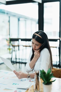 A portrait of a beautiful Asian female employee showing a stressed face while using the phone and financial documents on her desk.