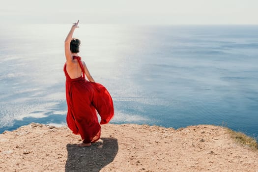 Side view a Young beautiful sensual woman in a red long dress posing on a rock high above the sea during sunrise. Girl on the nature on blue sky background. Fashion photo.