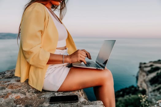 Successful business woman in yellow hat working on laptop by the sea. Pretty lady typing on computer at summer day outdoors. Freelance, travel and holidays concept.