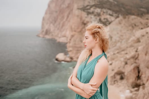 Side view a Young beautiful sensual woman in a mint long dress posing on a volcanic rock high above the sea during sunset. Girl on the nature on overcast sky background. Fashion photo
