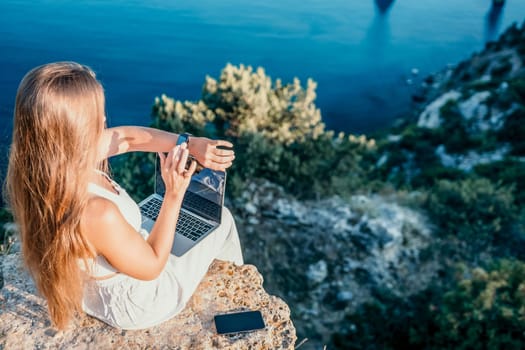 Successful business woman in yellow hat working on laptop by the sea. Pretty lady typing on computer at summer day outdoors. Freelance, travel and holidays concept.