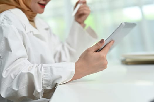 Cropped shot of smiling young muslim woman using digital tablet, enjoying freelance work in home office.