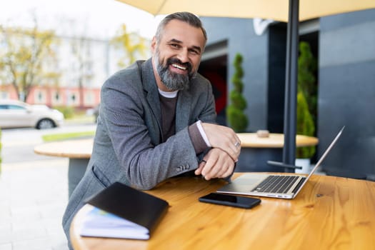 portrait of a mature adult male lawyer with gray hair and beard sitting on a terrace with a laptop.