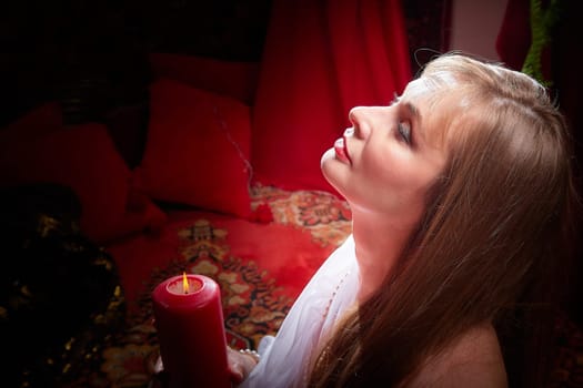 Beautiful arabian girl with candles in red room full of rich fabrics and carpets in sultan harem. Photo shoot of woman an oriental style odalisque. Model poses in sari as caring wife and hostess