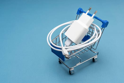 A phone charger in a supermarket cart on a blue background. Mini Shopping cart with Charger and Connecting Cable. side view from above