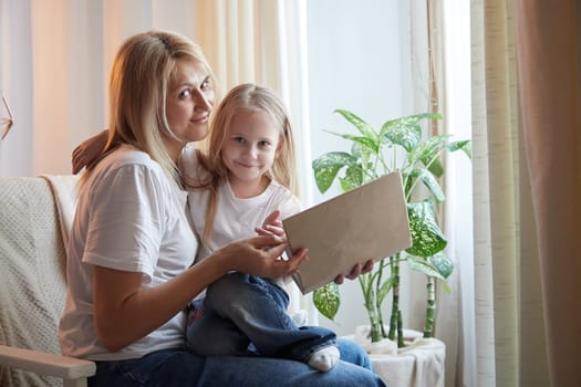 Happy loving family with mother and daughter reading book in living room. Woman mom and small child girl having convercation and studing inside of home