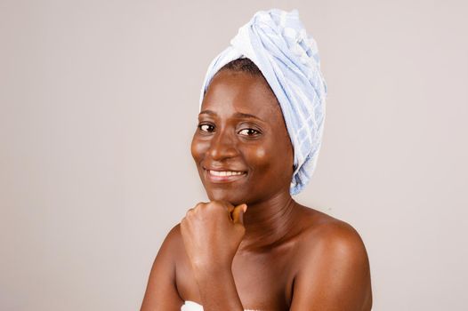 African girl sitting on gray background, bath towel knotted on the head smiling with the hand under the chin.