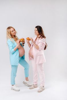 Two pregnant women in suits with hamburgers in their hands on a gray background.