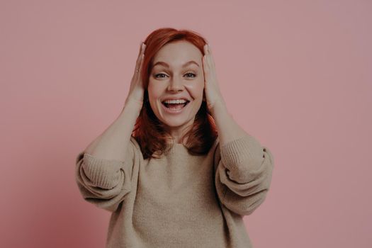 I cannot believe my eyes! Surprised emotional young female with red hair touching head with both hands and keeping mouth opened in excitement, dressed in beige jumper, isolated over pink background