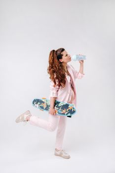A pregnant girl in a pink suit with a skateboard in her hands drinks juice on a gray background.