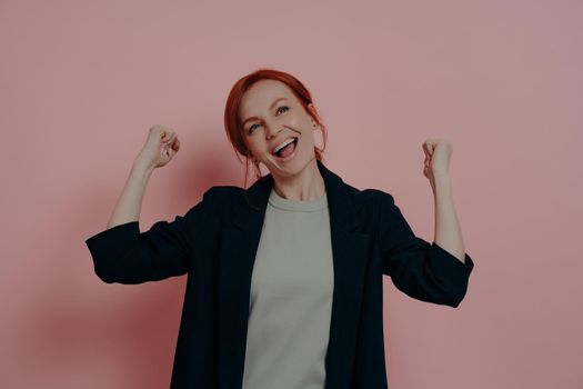 People and achievements. Indoor shot of young happy overjoyed ginger business woman raising hands in fists and celebrating triumph, screaming in excitement, isolated over pink studio background.